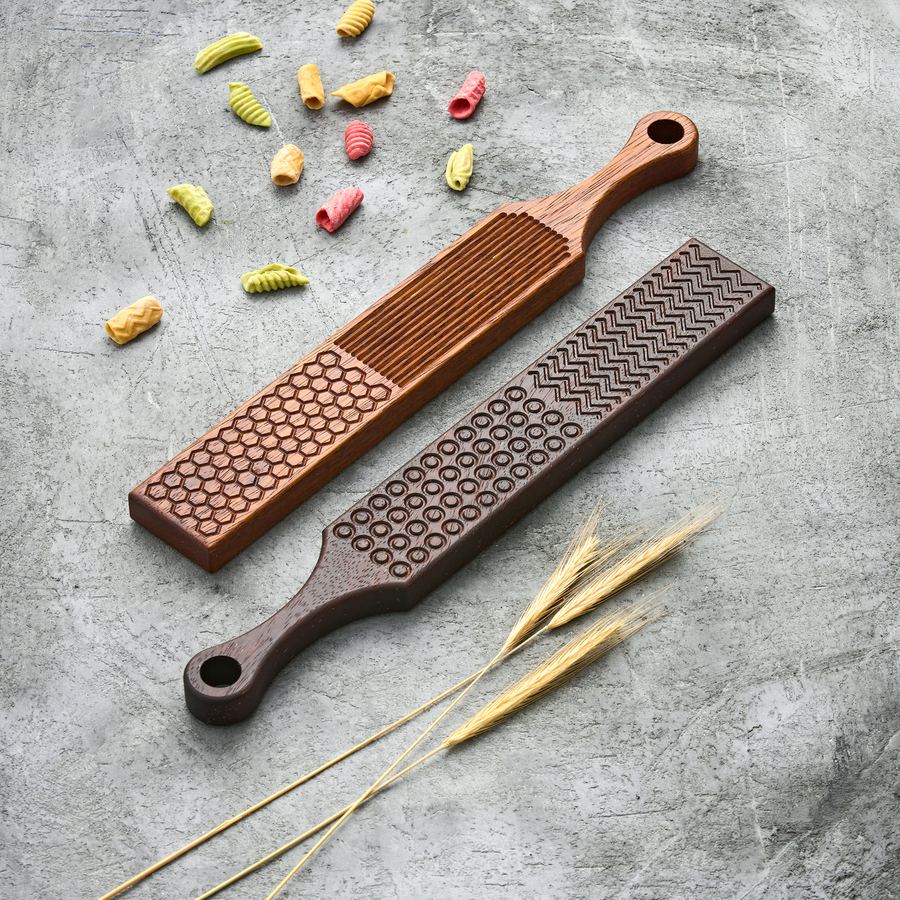 Two gnocchi boards with different groove patterns beside colored pasta and wheat stalks