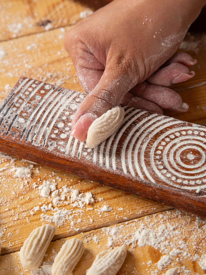 A piece of gnocchi dough rolling down a wooden ridged board, with shaped gnocchi beside it